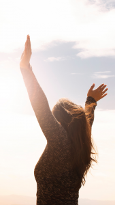 woman expressing freedom - arms raised high above her head, facing away from camera toward the shining sun